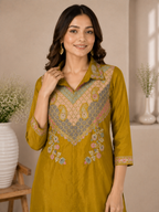 A woman wearing a mustard yellow kurta with intricate multicolor embroidery on the chest and cuffs, standing in a softly lit indoor setting with decorative vases.