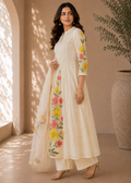 A woman models a cream-colored traditional Indian salwar kameez set with colorful floral embroidery on the sleeves and front drape, set against a warm beige background with soft window shadows.