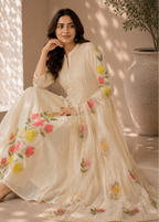 A woman models a cream-colored traditional outfit with vibrant pink, yellow, and green floral embroidery, posed in a sunlit indoor setting with a large textured vase in the background.