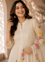 A woman smiling in a cream-colored, long-sleeved kurta with colorful floral embroidery in pink, yellow, and green, paired with matching sheer dupatta.