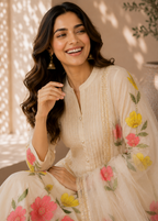A woman wearing a cream-colored traditional embroidered kurta set with pink and yellow floral patterns, seated in a warm, sunlit indoor setting.