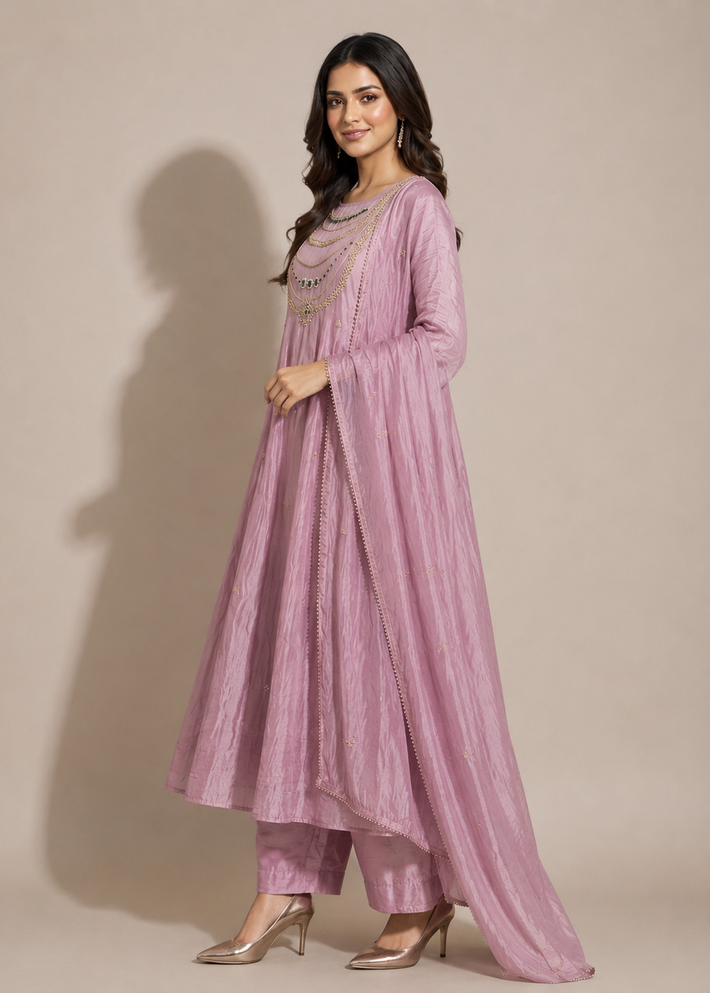 A woman modeling a dusty pink traditional Indian outfit consisting of a long kurta, matching trousers, and a draped dupatta, set against a solid beige studio background.