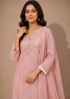 A woman modeling a light pink traditional outfit with a matching sheer dupatta, featuring small metallic embroidered details and white cuffs, set against a solid beige background.
