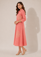 A woman modeling a coral pink long-sleeve midi dress with light beige floral embroidery on the chest and lower skirt, standing against a plain beige studio background.