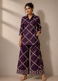 A woman modeling a matching purple two-piece kurta set with a white geometric lattice pattern, featuring a long-sleeve button-up top and wide-leg trousers, set against a neutral beige wall with soft window shadows.