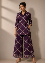 A woman modeling a matching purple two-piece kurta set with a white geometric lattice pattern, featuring a long-sleeve button-up top and wide-leg trousers, set against a neutral beige wall with soft window shadows.