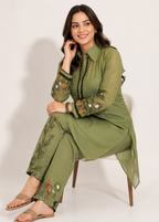 A woman wearing an olive green two-piece kurta set with floral embroidery on the sleeves and pant legs, sitting on a white chair against a light beige studio background.