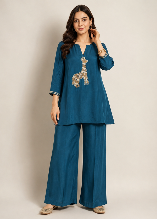A woman modeling a teal blue two-piece kurta and trouser set with a giraffe patchwork design on the chest, set against a plain beige studio background.