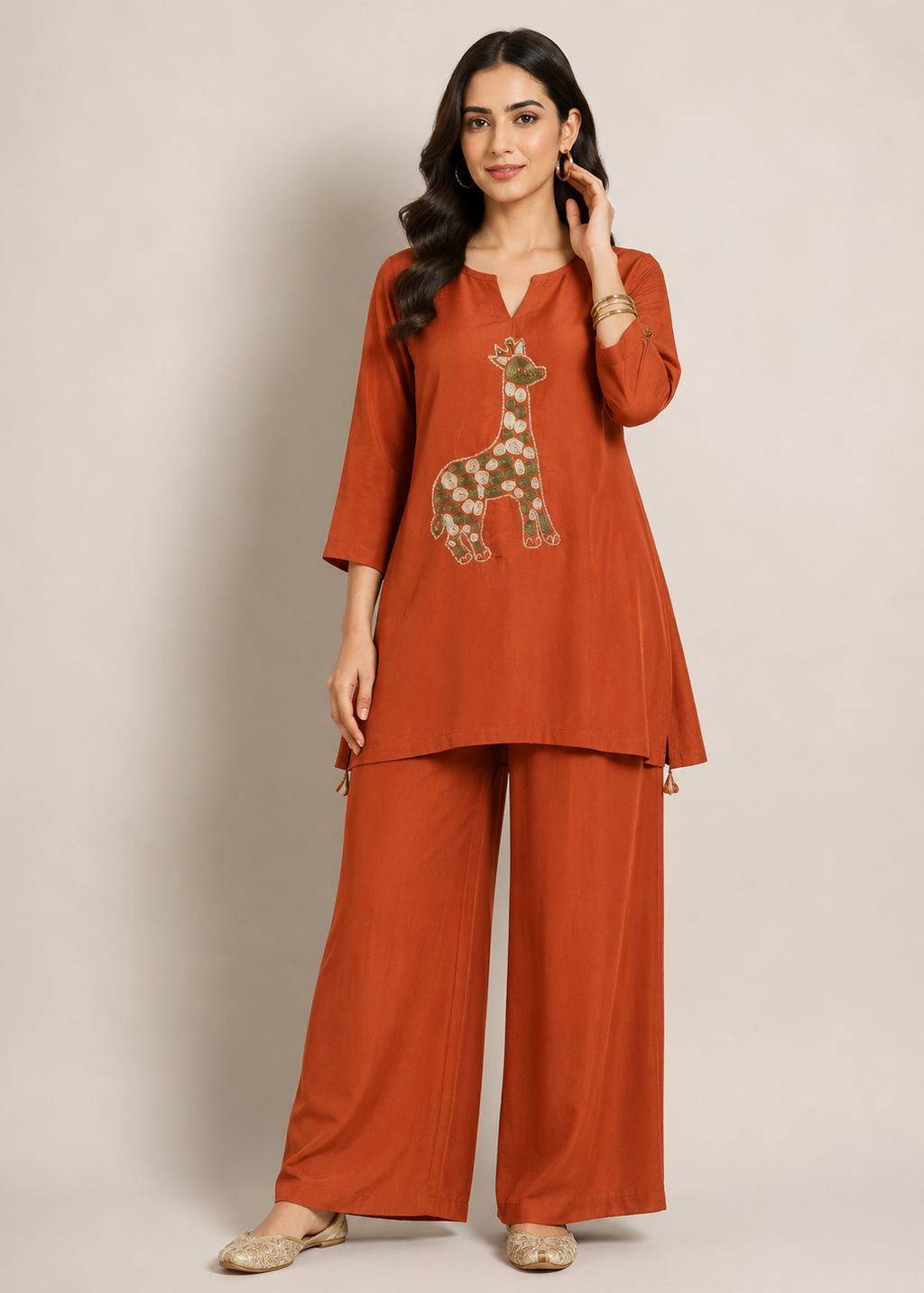 A woman modeling a rust-orange two-piece kurta and trouser set with a giraffe applique on the kurta, set against a plain beige studio background.