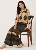A woman models a beige and black printed kurta set while sitting on a wooden chair against a plain light background.