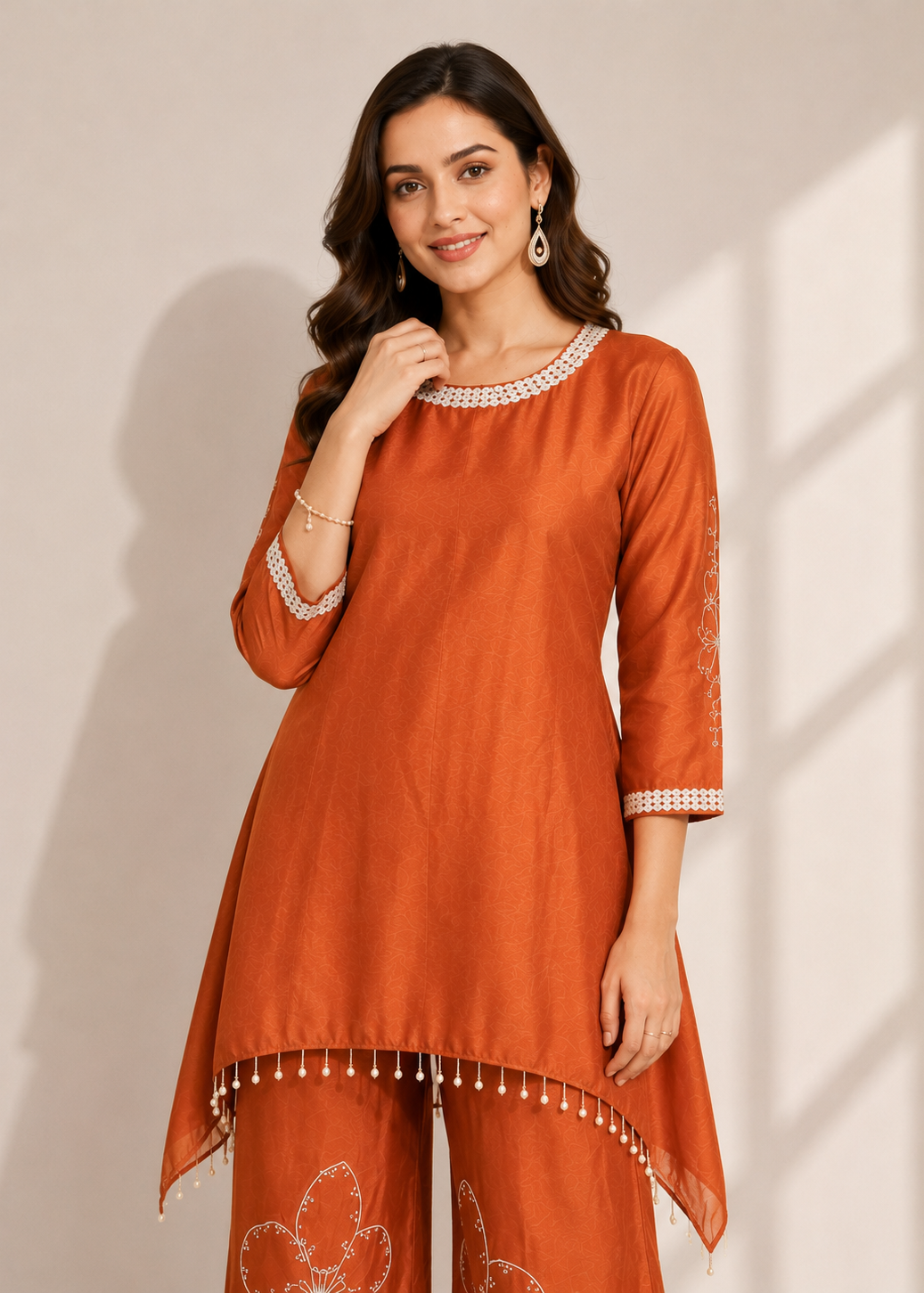 A woman modeling a rust-orange kurta and matching pants set with white embroidered details and beaded trim, set against a warm beige background with soft window shadow overlays.