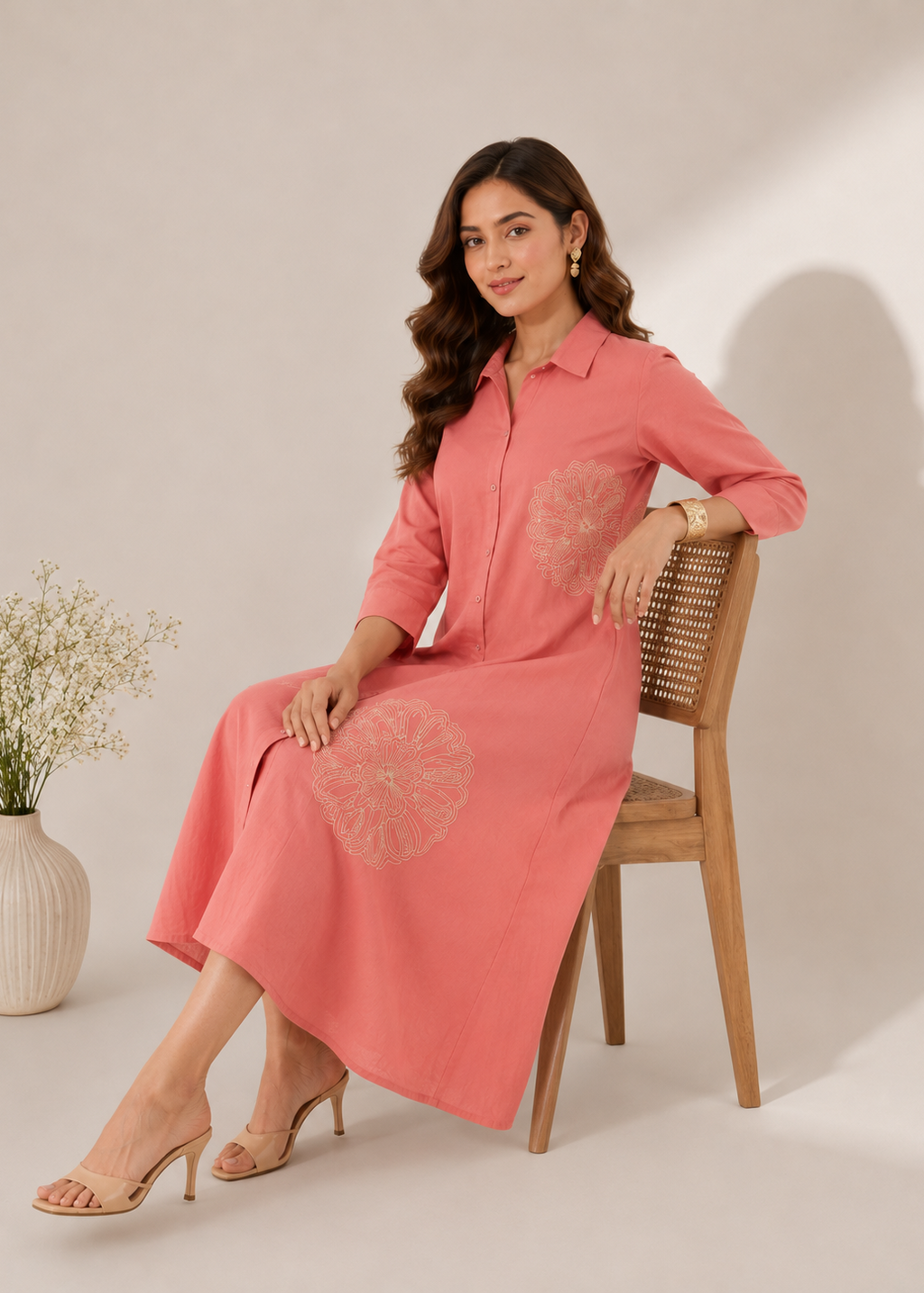 A woman wearing a pink long-sleeve midi dress with circular floral embroidery sits on a wooden chair in a studio setting.