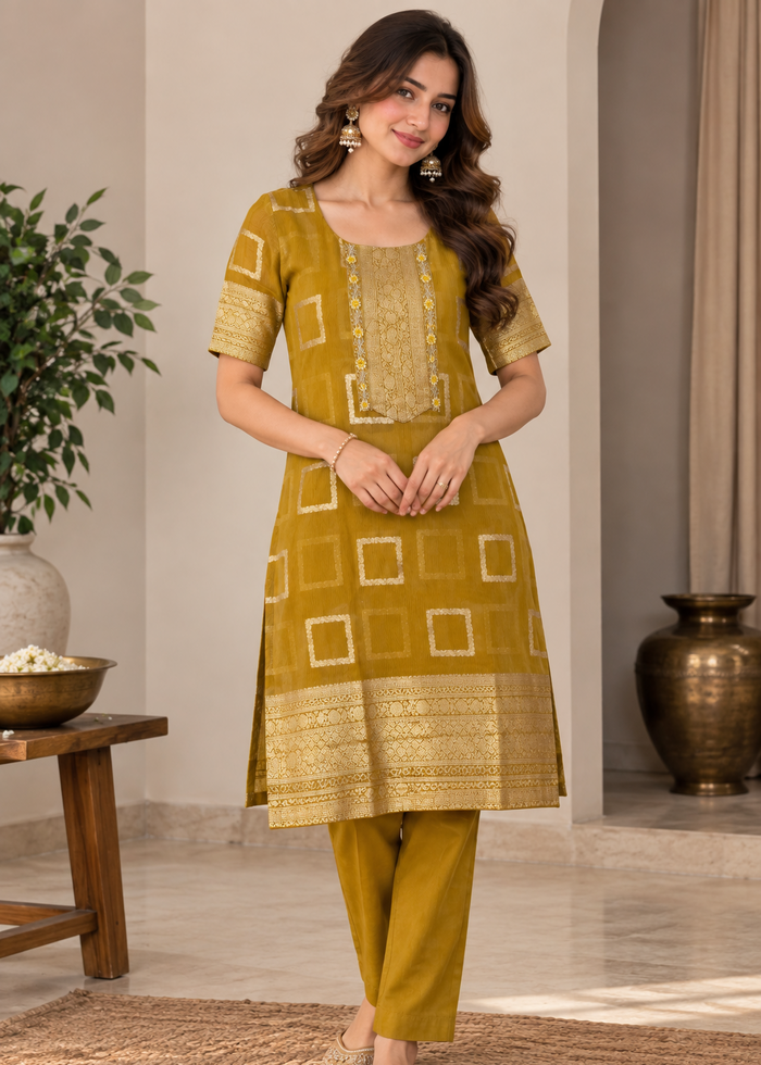 A woman modeling a mustard yellow ethnic kurta set with matching trousers in a styled indoor setting. The kurta features gold embroidery, geometric square patterns, and a decorative border, paired with matching solid trousers.