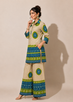 A woman modeling a beige two-piece kurta and trouser set with green and blue geometric patterns, standing against a plain beige background.