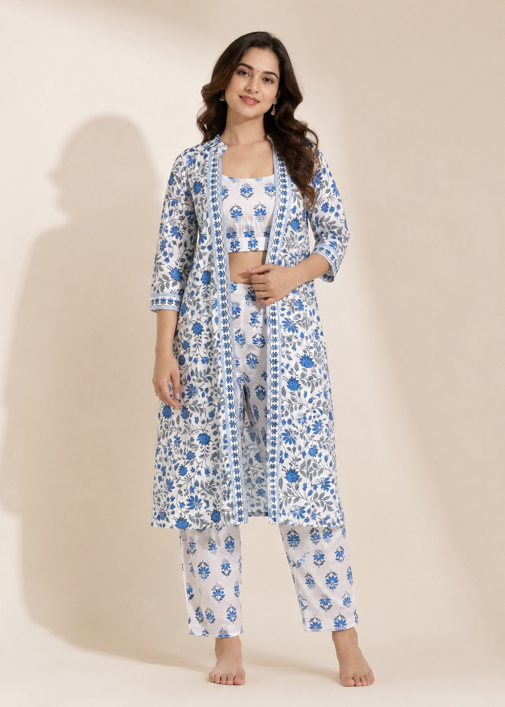 A woman modeling a three-piece printed loungewear set consisting of a white crop top, a matching open-front cardigan, and wide-leg pants, all featuring a blue floral and geometric block print pattern, set against a solid beige studio background.