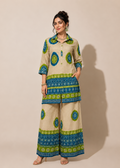 A woman modeling a beige two-piece kurta set with blue and green printed patterns, standing against a solid beige studio background.