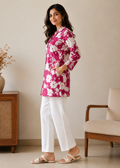 Woman wearing a pink floral top and white pants standing in a room with a chair and cabinet.