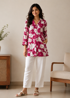 Woman wearing a pink floral blouse and white pants standing in a room with a wooden cabinet and chair.