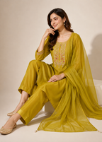 A woman models a three-piece olive green salwar kameez suit with a matching sheer dupatta. The kurta features gold and silver embroidery on the chest, and she is seated on a white block against a warm beige studio background.