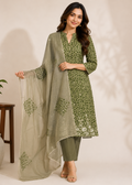A woman models a three-piece Indian ethnic suit set featuring an olive green printed kurta, matching olive green pants, and a sheer sage green dupatta with printed motifs, set against a warm beige background.