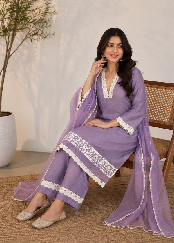 A woman models a lavender purple ethnic suit set with white lace trim, consisting of a kurta, trousers, and a matching dupatta, while sitting on a wooden chair.