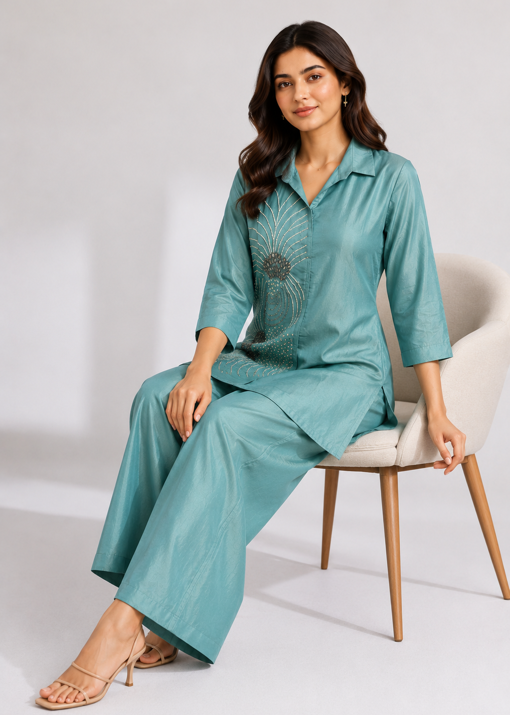 A woman models a matching two-piece loungewear set in a sage green/teal color, sitting on a light beige armchair. The set includes a short-sleeve collared shirt with a subtle embroidered sunburst pattern on the chest and matching wide-leg trousers.
