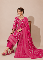 A woman seated on a wooden chair wearing a bright pink traditional outfit with white floral and geometric patterns, matching pants, and a long draped dupatta, against a plain beige studio background.
