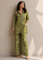 A woman models a matching two-piece pajama set with a green and white abstract floral pattern, consisting of a long-sleeve button-up shirt and wide-leg pants, set against a neutral beige wall with soft window shadows.