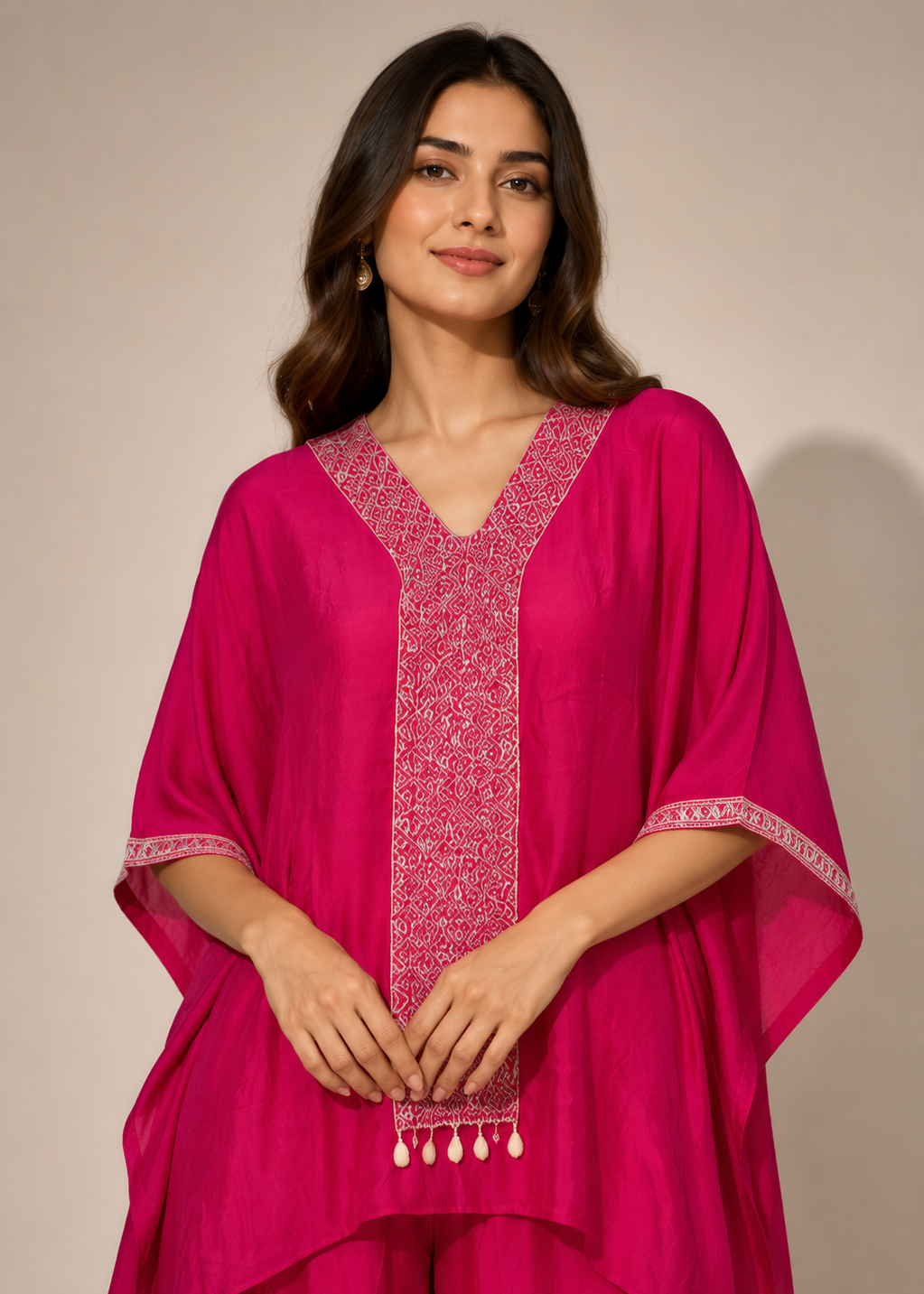 A woman modeling a bright magenta pink kaftan-style tunic with intricate white embroidery along the V-neckline, sleeve cuffs, and hem, paired with matching trousers against a plain beige studio background.