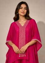 A woman modeling a bright magenta pink kaftan-style tunic with intricate white embroidery along the V-neckline, sleeve cuffs, and hem, paired with matching trousers against a plain beige studio background.