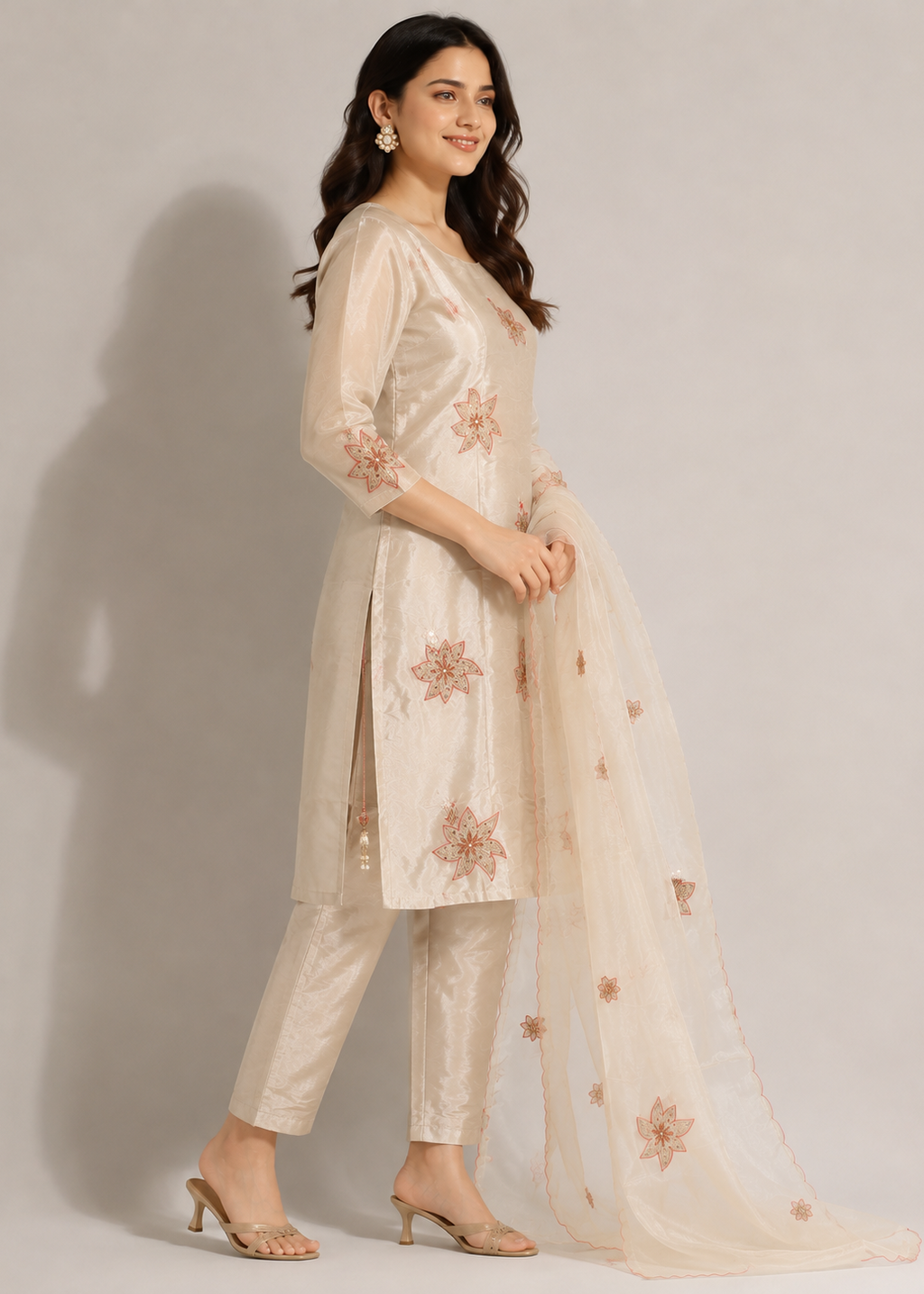 A woman modeling a traditional Indian salwar kameez suit in a light beige/cream color with peach embroidered floral patterns, paired with matching trousers and a sheer dupatta, set against a plain light gray studio background.
