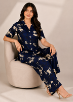 A woman models a navy blue short-sleeve button-up shirt and matching wide-leg trousers set with a beige floral print, seated on a modern beige armchair.