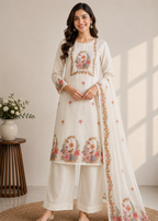 A woman models a traditional Indian salwar kameez suit set in off-white with intricate floral embroidery in pink, orange, and green. The set includes a long tunic (kameez), matching wide-leg pants (salwar), and a sheer dupatta.