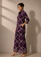 A woman modeling a deep purple two-piece kurta set with a white geometric lattice pattern, standing against a warm beige wall with soft window shadows.