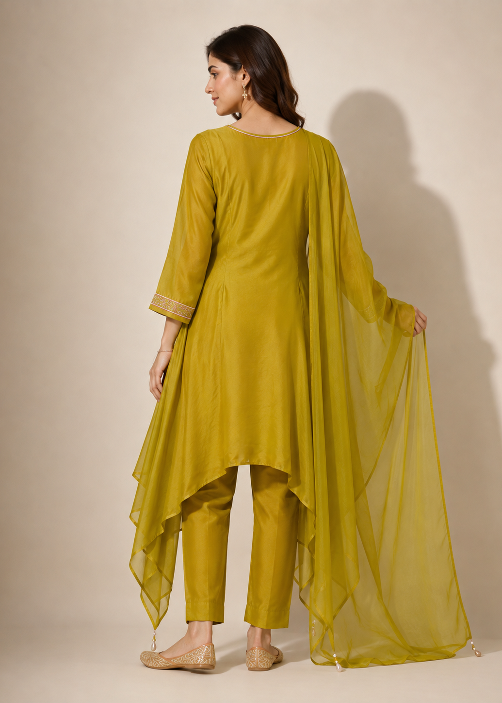A woman modeling a three-piece olive green traditional outfit with a high-low draped kurta, matching trousers, and a sheer dupatta, shown from the back against a warm beige studio background.