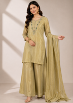 A woman models a traditional Indian salwar kameez suit in a light olive green or khaki-gold color, featuring a long kurta with embroidered neckline, matching wide-leg trousers, and a draped dupatta, set against a neutral studio backdrop with soft window shadow projections.
