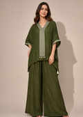 A woman modeling a dark olive green two-piece outfit consisting of a loose, short-sleeved V-neck tunic with light green embroidered trim and matching wide-leg trousers, set against a plain beige studio background.