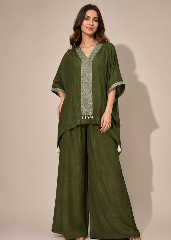 A woman modeling a dark olive green two-piece outfit consisting of a loose, short-sleeved V-neck tunic with light green embroidered trim and matching wide-leg trousers, set against a plain beige studio background.