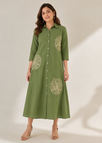 A woman modeling a green button-front midi dress with three-quarter sleeves and large beige floral embroidery on the chest and skirt, photographed against a plain light beige studio background.