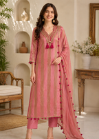 A woman models a three-piece pink ethnic suit set featuring a kurta with gold vertical stripes, matching trousers, and a sheer dupatta with pom-pom trim, set against a warm, decorated interior background.