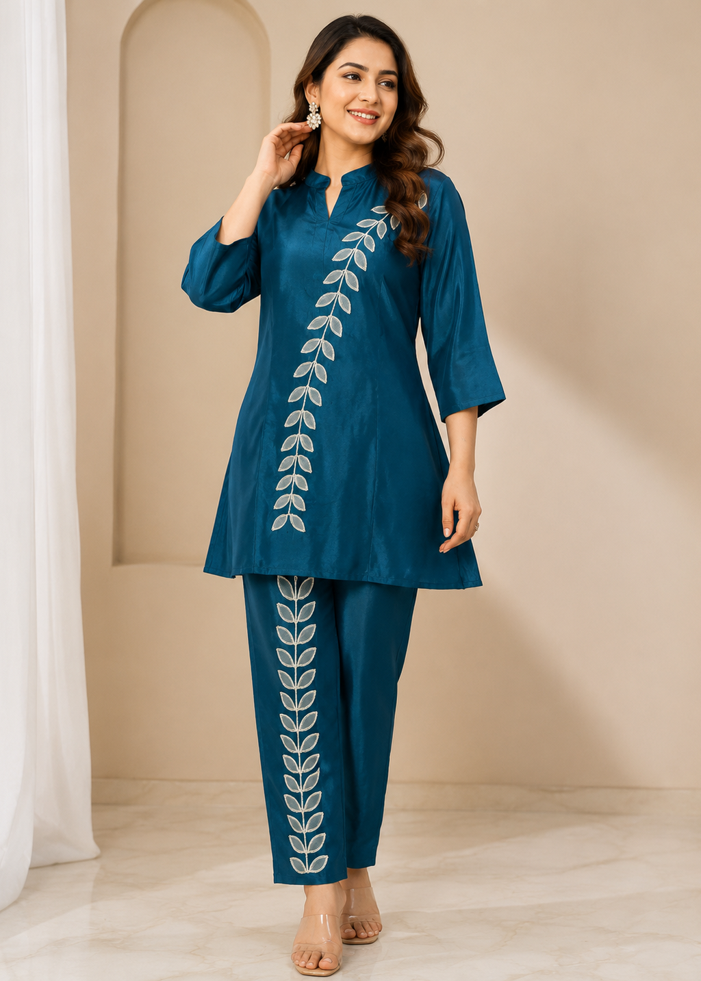 A woman modeling a teal blue kurta and trouser set with white leaf embroidery down the front, standing in a warm beige studio setting.