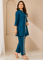 A woman modeling a teal blue two-piece kurta set with white leaf embroidery down the front and trouser leg, standing in a beige arched room setting.