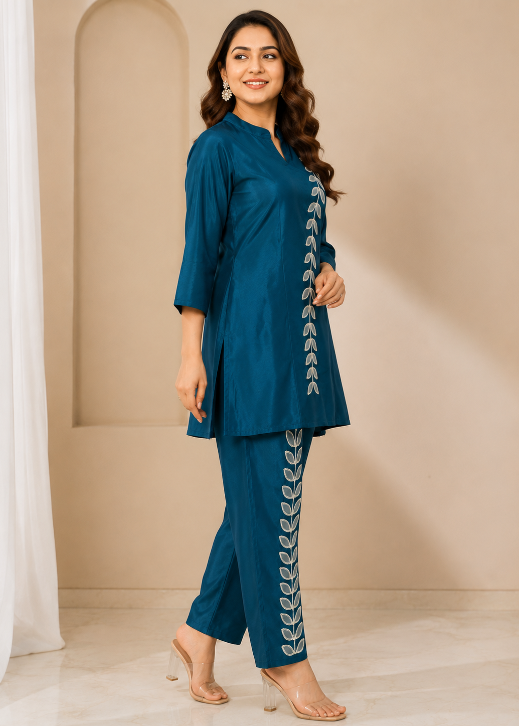 A woman modeling a teal blue two-piece kurta set with white leaf embroidery down the front and trouser leg, standing in a beige arched room setting.