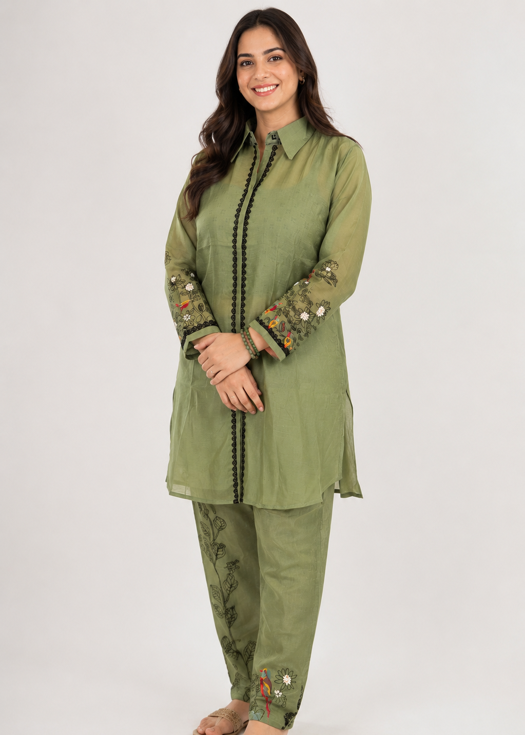 A woman modeling an olive green traditional kurta set with matching pants, featuring black embroidered trim down the front and sleeves, and multicolor floral embroidery on the cuffs and hem, set against a plain light gray background.