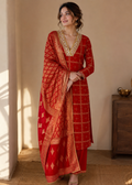 A woman models a traditional red Indian salwar kameez suit with gold zari embroidery and a matching red dupatta, set against a warm beige wall and wooden furniture.