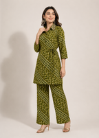 A woman modeling a matching olive green printed kurta and trouser set with a tie waist belt, photographed against a plain light beige studio background.