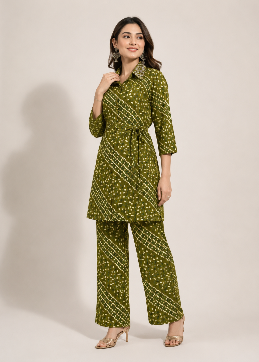 A woman modeling a matching olive green printed kurta and trouser set with a tie waist belt, photographed against a plain light beige studio background.