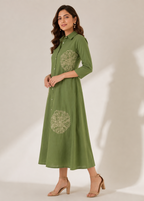 A woman modeling a green midi shirt dress with three-quarter sleeves and circular floral embroidery on the chest and lower skirt, paired with beige heeled sandals against a warm beige studio backdrop.