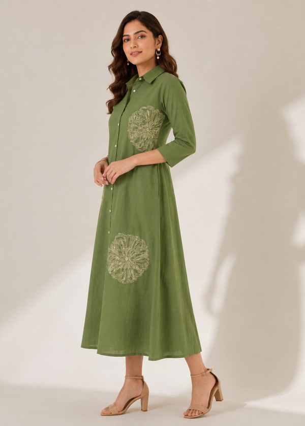 A woman modeling a green midi shirt dress with three-quarter sleeves and circular floral embroidery on the chest and lower skirt, paired with beige heeled sandals against a warm beige studio backdrop.