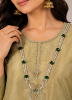 A close-up of a woman wearing an ornate gold-toned traditional necklace with multiple strands of white stones and green gemstone accents, paired with a matching olive green embroidered top.